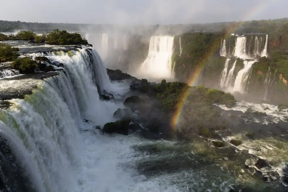 Iguazu Falls at sunrise, with mist rising above the cascades and a rainbow forming over the water