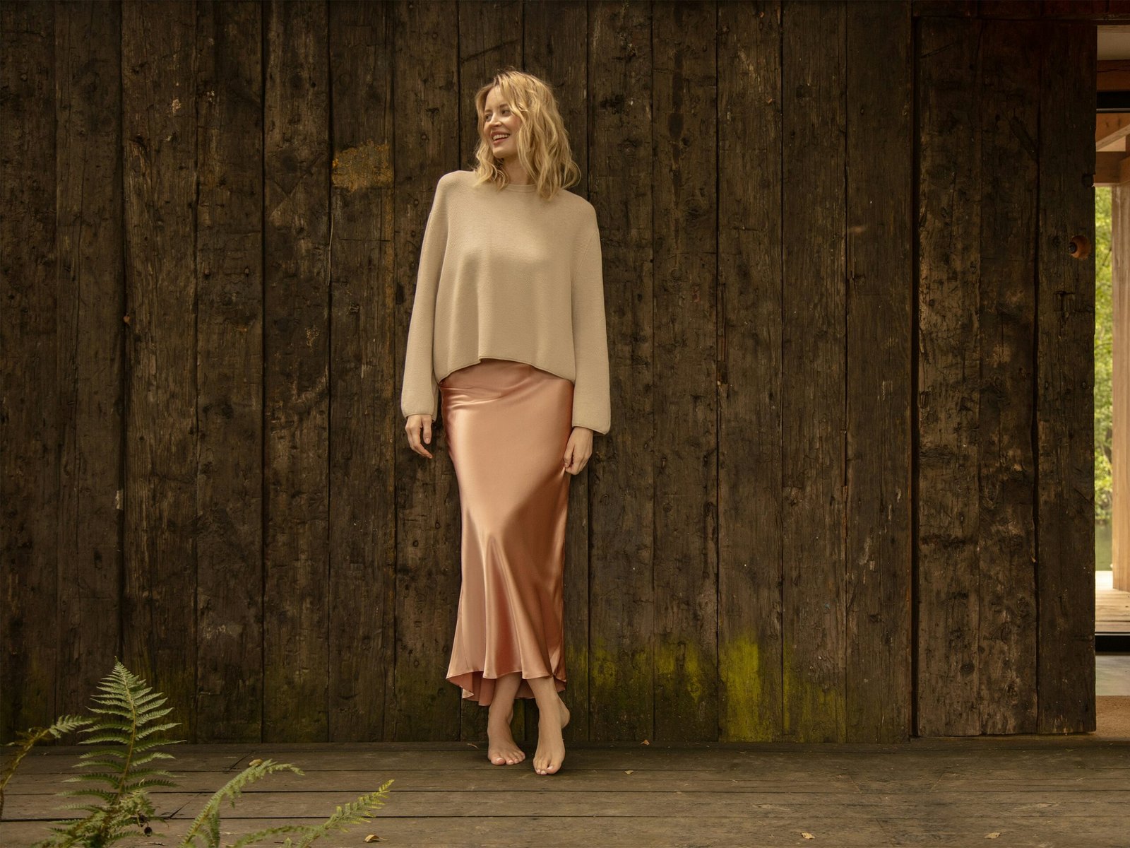 Woman wearing a blush silk skirt and beige top in front of a dark wooden wall